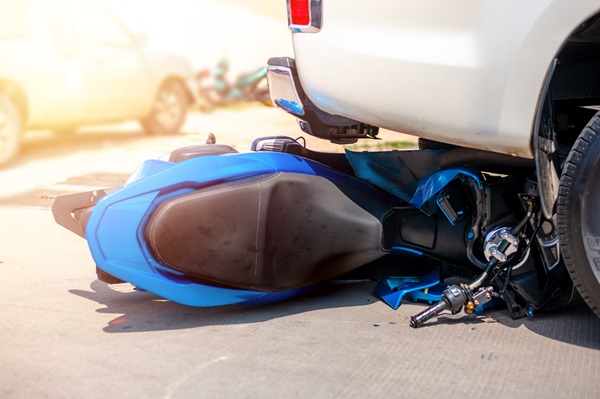 Blue motorcycle lying on its side wedged under the rear of a white pickup truck after a collision.