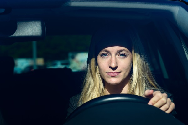 Young woman with long blonde hair sitting in the driver’s seat of a car, looking forward with one hand on the steering wheel.