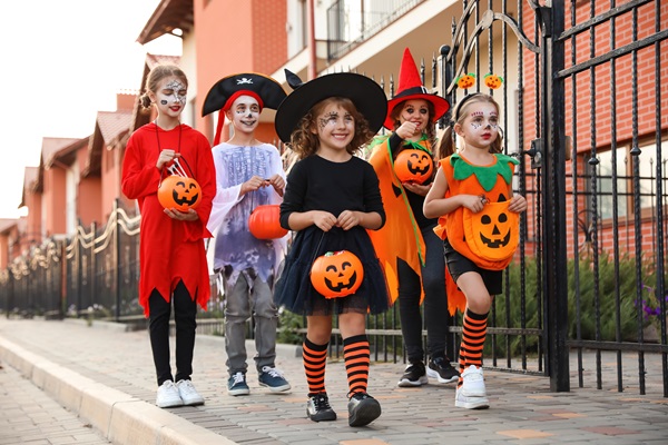 Group of children in Halloween costumes walking on a neighborhood sidewalk while carrying pumpkin candy buckets.