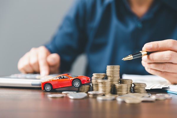 Person calculating money with stacked coins and a small red toy car on a desk, symbolizing auto insurance refunds or car-related expenses.