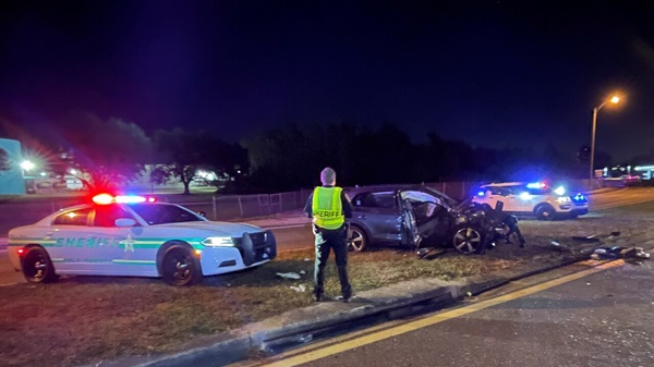 A severely damaged dark SUV after a nighttime head-on collision in Lakeland, FL, surrounded by two Sheriff patrol cars with flashing lights and an officer standing in the foreground.