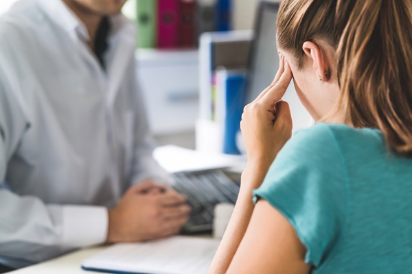 A woman rubbing her temples in distress while sitting across from a doctor in a medical office, illustrating the consultation for persistent brain injury symptoms or chronic headaches after a Tampa car accident.