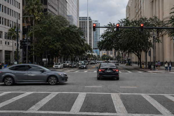 A black sedan runs a red light at a busy city intersection as a second car enters from a cross street.
