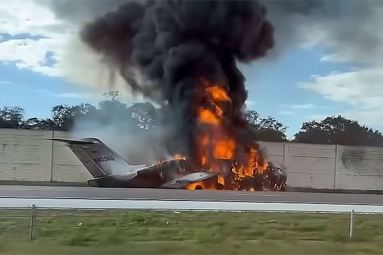 A small private plane engulfed in flames on a runway with heavy black smoke, illustrating the fire and burn hazards small plane crash victims can face.
