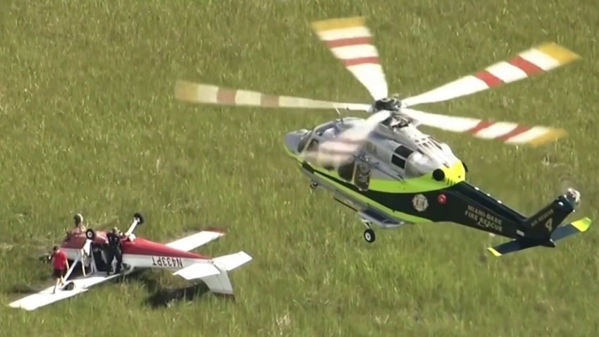 A Miami-Dade Fire Rescue helicopter approaches a small red and white private plane that made an emergency landing in a grass field, showing first responders arriving at a general aviation accident.
