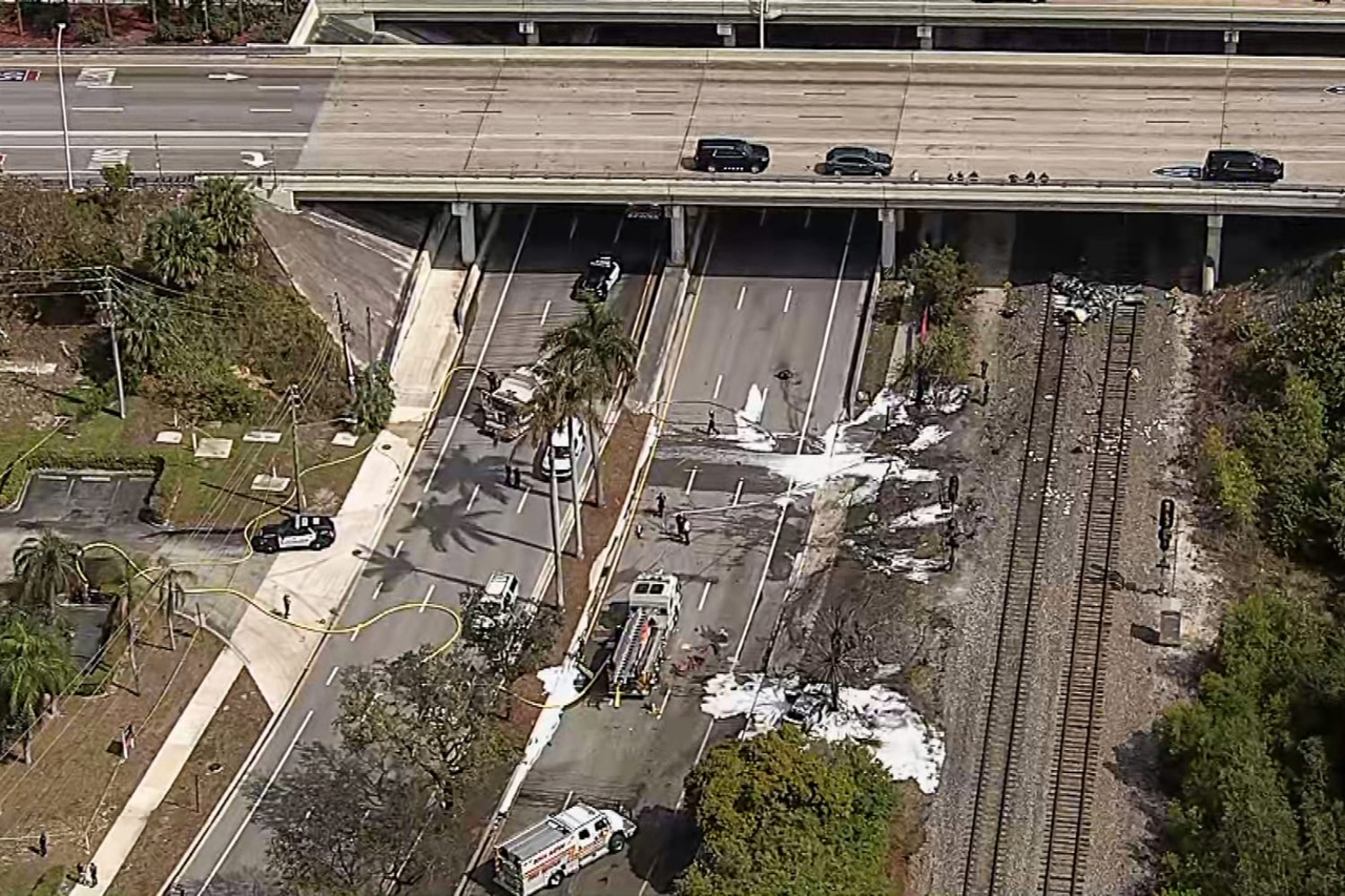 Aerial view of a highway underpass and railroad tracks covered in firefighting foam after a small plane crash, with fire trucks and police vehicles on scene.