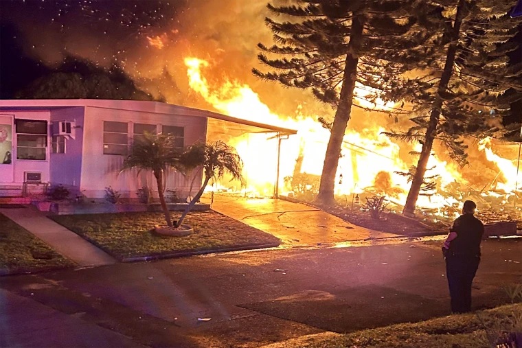 A police officer stands near a mobile home community at night as flames from a small plane crash engulf a nearby structure, showing the risk aviation accidents pose to residents on the ground.